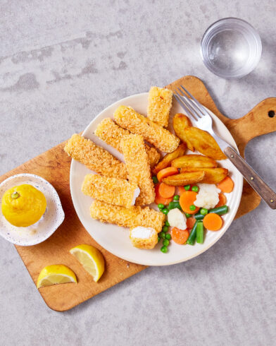 Ein Teller mit FRoSTA Cornflakes-Stäbchen, FRoSTA Kartoffelecken, FRoSTA Mischgemüse und einer Gabel auf Holz. Zitrone und Wasser daneben.