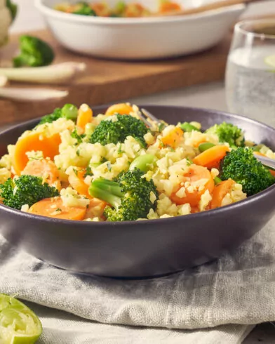 Eine ASIAN STYLE BOWL mit FRoSTA Tiefkühlreis, Brokkoli, Karotten & Gemüse steht auf einer Serviette. Im Hintergrund frische Zutaten.