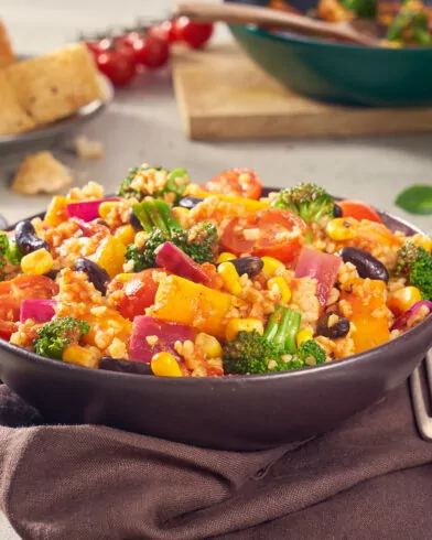 Eine farbenfrohe FRoSTA Mexican Style Bowl mit Mais, Brokkoli, Bohnen, Tomaten und roten Zwiebeln steht auf einer Serviette. Brot und ein Zitronengetränk runden das Menü ab.