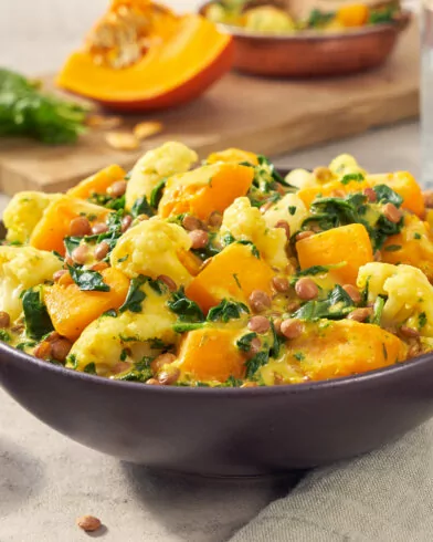 Eine indische Schüssel mit FRoSTA-Tiefkühlgemüse-Curry aus Blumenkohl, Kürbis, Spinat und Linsen steht auf dem Tisch mit Besteck und Gewürzen.