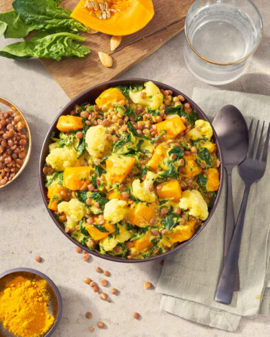 Eine FRoSTA Indian Style Bowl mit Curry, Blumenkohl, Kürbis, Spinat und Linsen steht neben Besteck auf einer Serviette.