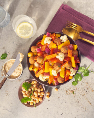 Eine lebendige Falafel-Bowl mit FRoSTA Tiefkühlprodukten, frischem Salat, goldenem Besteck, Zitronenwasser, Hummus, Kichererbsen und Koriander auf einer hellen Unterlage.