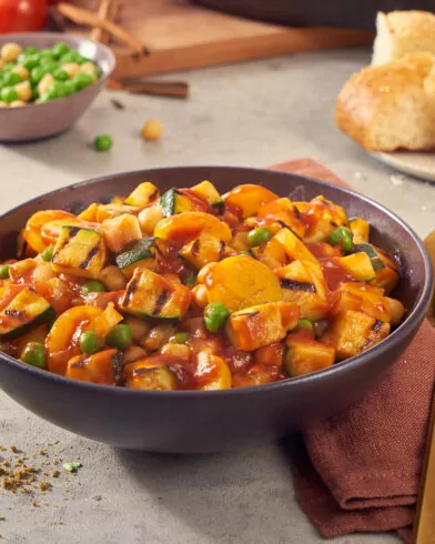 Eine Schüssel FRoSTA-Gemüsecurry mit Erbsen, Karotten, Zucchini und Kichererbsen in roter Sauce, serviert mit Brot und Koriander.