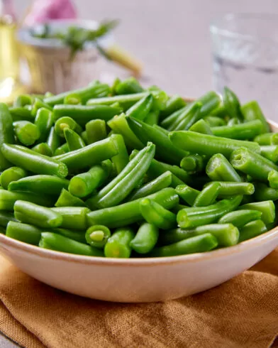 Eine Schüssel mit frischen, gekochten FRoSTA Tiefkühlprodukten Bioland Junge Brechbohnen steht auf einer Serviette, dazu ein Glas Wasser und etwas Öl.