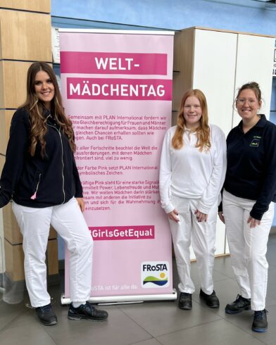 Drei Frauen in weißen Uniformen lächeln neben einem rosa Weltmädchentag-Banner über die Rechte von Mädchen, in der Nähe stehen FRoSTA-Tiefkühlprodukte. #GirlsGetEqual.