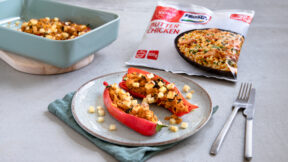 Ein Teller mit zwei gefüllten Paprikaschotenhälften gefüllt mit Gemüse und Kartoffelwürfeln, dazu im Hintergrund eine Packung FROSTA Butter Chicken.