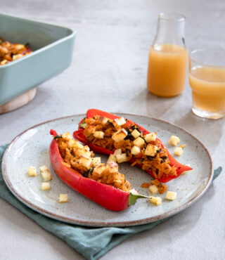Ein Teller mit zwei gefüllten Paprikaschoten, hergestellt aus FRoSTA Tiefkühlprodukten, garniert mit Käse, steht neben einem Glas und einer Kanne Saft.