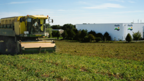 Ein gelb-grüner Mähdrescher wirbt für Nachhaltigkeit, während er in der Nähe eines weißen Industriegebäudes mit der Aufschrift „FRoSTA“, einem Hersteller von Tiefkühlprodukten, arbeitet.