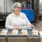 Eine Frau mit Haube, Brille und weißer Uniform hält FRoSTA Tiefkühl-Fischstäbchen an einem nachhaltigen Förderband mit weiteren FRoSTA Paketen.