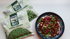 Zwei Beutel FRoSTA Bio-Tiefkühlgemüse (Erbsen, Bohnen) liegen neben einer Schale Erbsen-Bohnen-Salat mit roten Zwiebeln, Granatapfel und Nüssen.