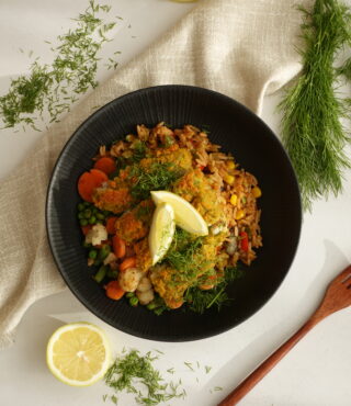 Eine schwarze Schüssel mit FRoSTA Bratreis-Bowl, Gemüse und Kräutern, garniert mit Zitronenspalten und frischem Dill auf hellem Tuch.