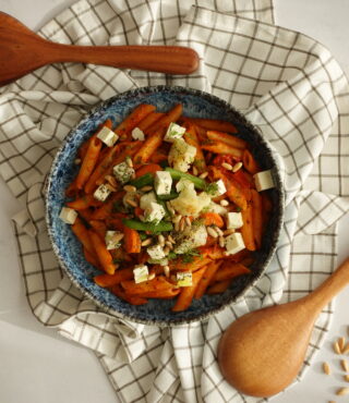 Eine blaue Schüssel Penne all’Arrabbiata von FRoSTA mit roter Sauce, Kräutern, Käse und Nüssen liegt auf einem Tuch mit Holzutensilien.