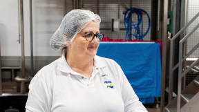 Eine Frau in Brille, Haarnetz und Polo zeigt bei FRoSTA-Tiefkühlprodukten im Betrieb Engagement und Professionalität.