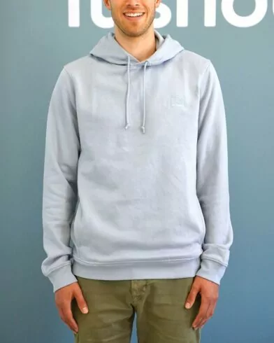 Eine Person in einem hellgrauen FRoSTA Hoodie Unisex mit olivgrüner Hose steht lächelnd vor einem blauen Hintergrund mit weißem Text, die Hände entspannt an den Seiten.