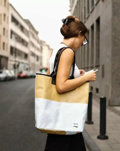 Eine Frau mit hochgestecktem Haar geht eine Straße entlang, hält ihr Telefon in der Hand, trägt eine Brille und trägt eine große FRoSTA-Einkaufstasche.