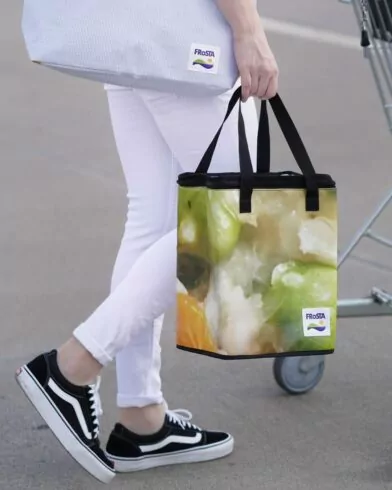 Eine Person in weißer Hose und schwarzen Turnschuhen steht im Freien bei einem Einkaufswagen und hält eine FRoSTA Tiefkühltasche mit FRoSTA Tiefkühlprodukten in der Hand.