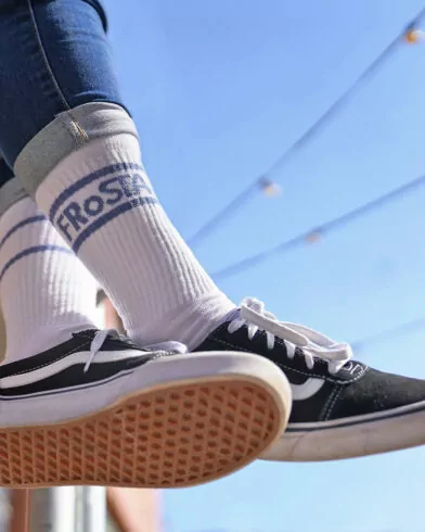 Person trägt FRoSTA Socken Weiß-Rot mit „FROST“ Schrift, hochgekrempelte Jeans und Sneaker – draußen unter Lichterkette und blauem Himmel.
