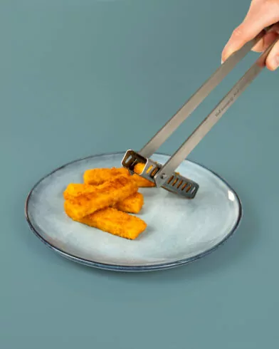 Eine Hand benutzte eine Wendezange, um panierte Fischstäbchen von FRoSTA von einem blauen Keramikteller aufzunehmen oder darauf abzulegen. Der Teller steht vor einem schlichten, hellblauen Hintergrund.