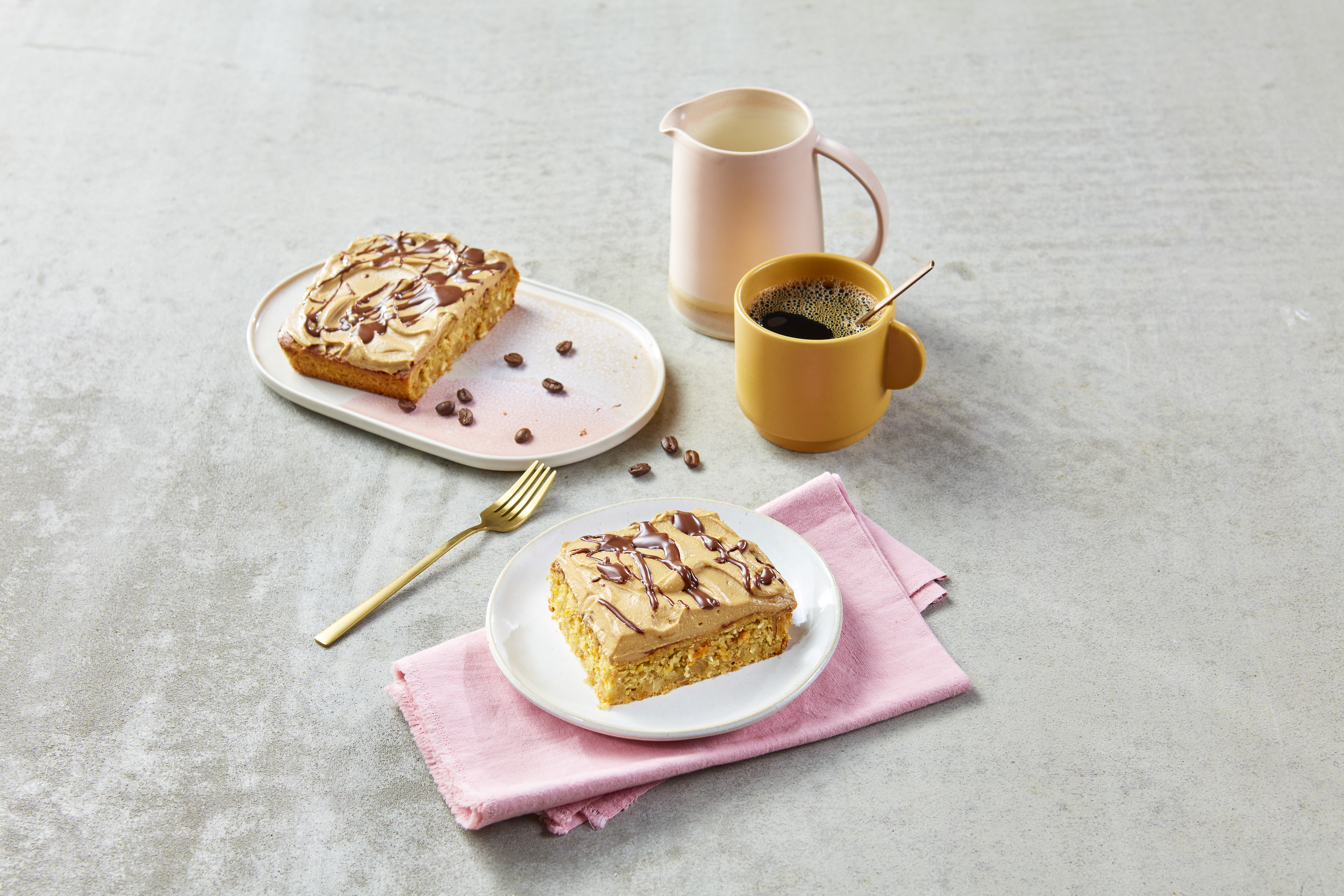 Ein Stück FRoSTA Tiefkühlprodukt-Kuchen mit Espresso-Buttercreme und Schokolade auf weißem Teller, daneben Kaffee und Deko.