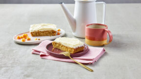 Ein Stück Karottenkuchen mit Frischkäse-Frosting und Mango von FRoSTA auf Tellern, dazu Besteck, Tasse und Teekanne auf grauer Fläche.