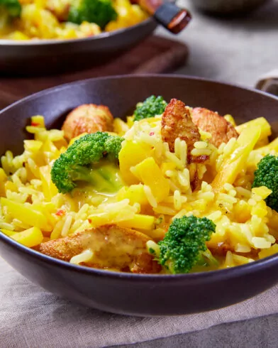 Eine Schüssel mit gelbem FRoSTA-Reis mit Brokkoli und Hähnchenfleisch, gewürzt mit Curry, auf einem grauen Tuch; weitere Schüsseln mit Lebensmitteln im Hintergrund.