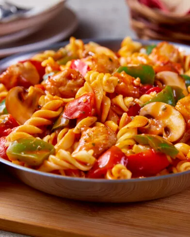 Eine FRoSTA Hähnchen Gulasch Pfanne mit bunter Pasta, Gemüse und Hähnchen in Tomatensauce steht auf einem Brett, Brot im Hintergrund.