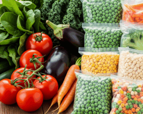 Frisches Gemüse - Tomaten, Karotten, Spinat, Grünkohl, Gurken, Auberginen - neben mehreren Tüten FRoSTA-Tiefkühlgemüse auf einer Holzfläche.