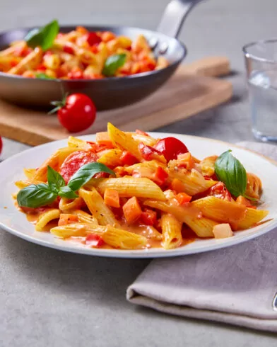 Eine FRoSTA Nudel Rahm Pfanne und Penne mit Soße, Basilikum, Gemüse neben Brot, Wasser, Tomaten, Gabel und Serviette.