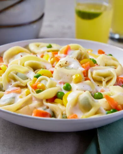 Tortellini in weißer Soße mit FRoSTA Sommergemüse (Karotten, Erbsen, Mais), dazu grüne Serviette und zwei Gläser gelber Drink.