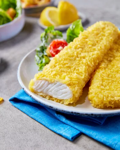 Ein Teller mit zwei FRoSTA Schlemmerfilet Fischfilets in goldgelber Panade, eines davon aufgeschnitten, sodass das weiße Fischinnere sichtbar ist. Daneben frischer grüner Salat mit einer roten Tomatenhälfte. Im Hintergrund unscharf ein Stück Zitrone und eine Schale mit weiteren Salatstücken.