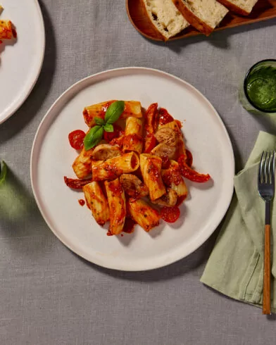 Ansicht von oben: Tisch mit FRoSTA Hähnchen alla Caprese, Rigatoni, grünen Servietten, Wasser und Brotscheiben auf grauer Decke.