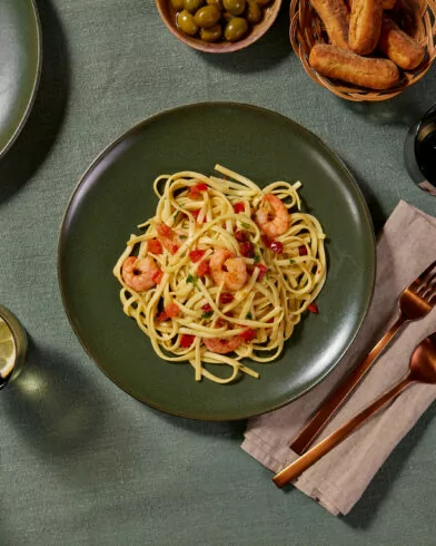 Linguine mit FRoSTA Garnelen, roten Paprika, Besteck auf grünem Tisch; dazu Zitrone, Oliven, Brotsticks und weiteres Pastagericht.