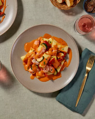 Zwei Teller FRoSTA Wildlachs und Garnelen mit Rigatoni, Gemüse, rosa Getränken, Brotkorb und Gewürzen auf beigem Tischtuch.