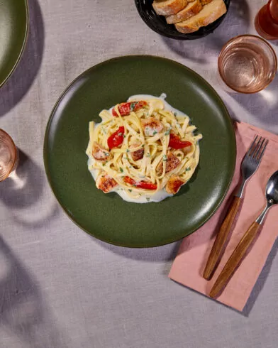 Draufsicht auf einen Tisch mit cremigen Caesar-Chicken-Spaghetti nach FRoSTA-Art, Tomatenwürfeln, rosa Servietten, Brot und Gläsern.