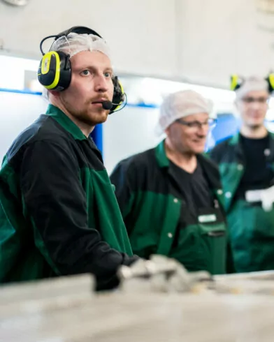Drei Arbeiter verarbeiten FRoSTA-Tiefkühlprodukte in Schutzkleidung an Industrieanlagen. Einer trägt Headset, Hintergrund unscharf.