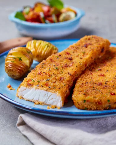 Zwei knusprige FRoSTA Backofen Fisch Mediterraner Art Filets mit Hasselback-Kartoffeln, Gemüse im Hintergrund, Glas Wasser daneben.