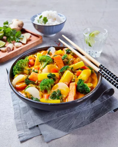 Eine Schüssel FRoSTA gelbes Gemüsecurry mit Brokkoli, Karotten & mehr, Stäbchen. Reis, Wasser mit Limette und Kräutern im Hintergrund.