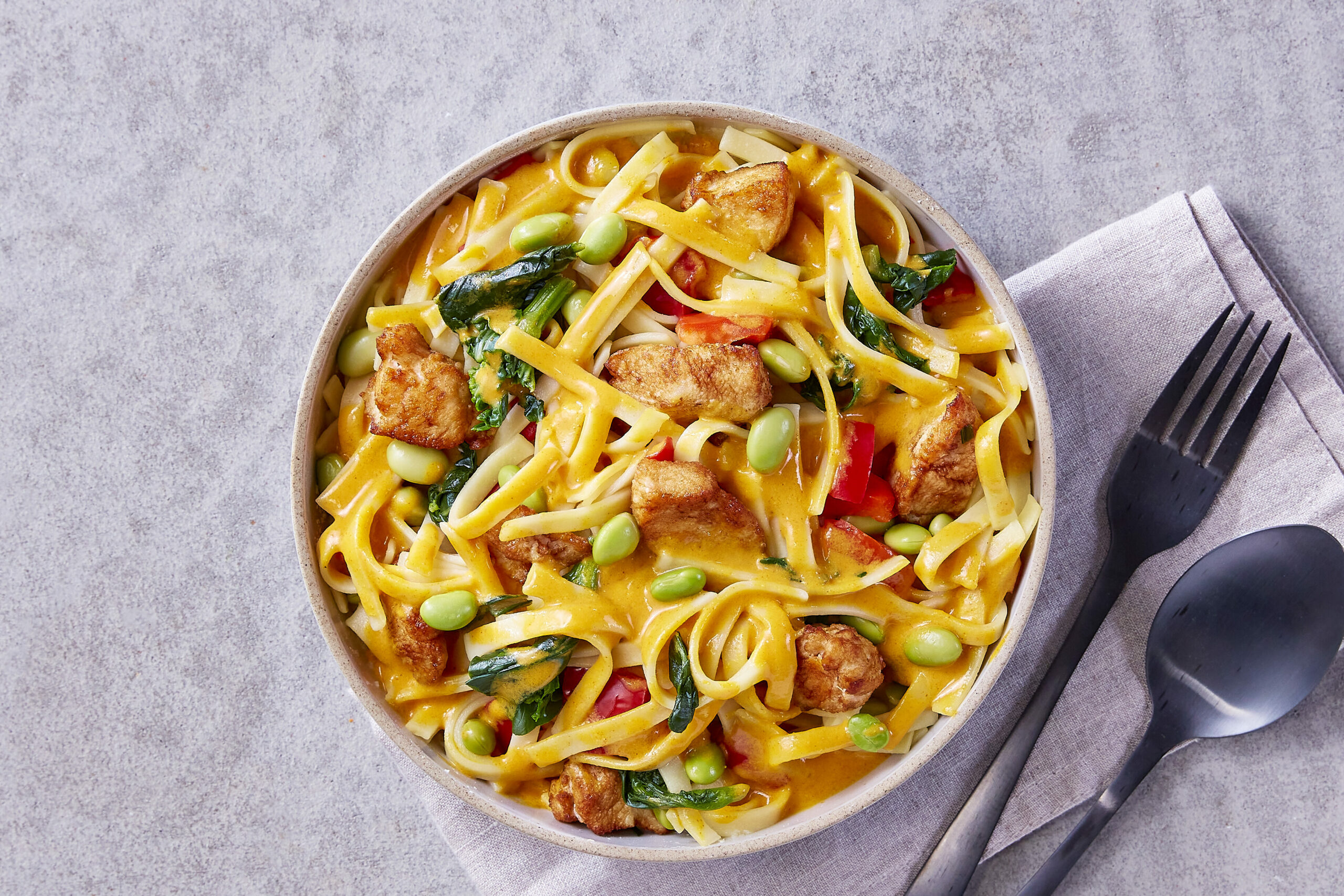 Eine Schüssel FRoSTA-Nudelpfanne mit Tofu, Edamame, roter Paprika und Blattgemüse in Soße, serviert auf einer Serviette mit Gabel und Löffel.