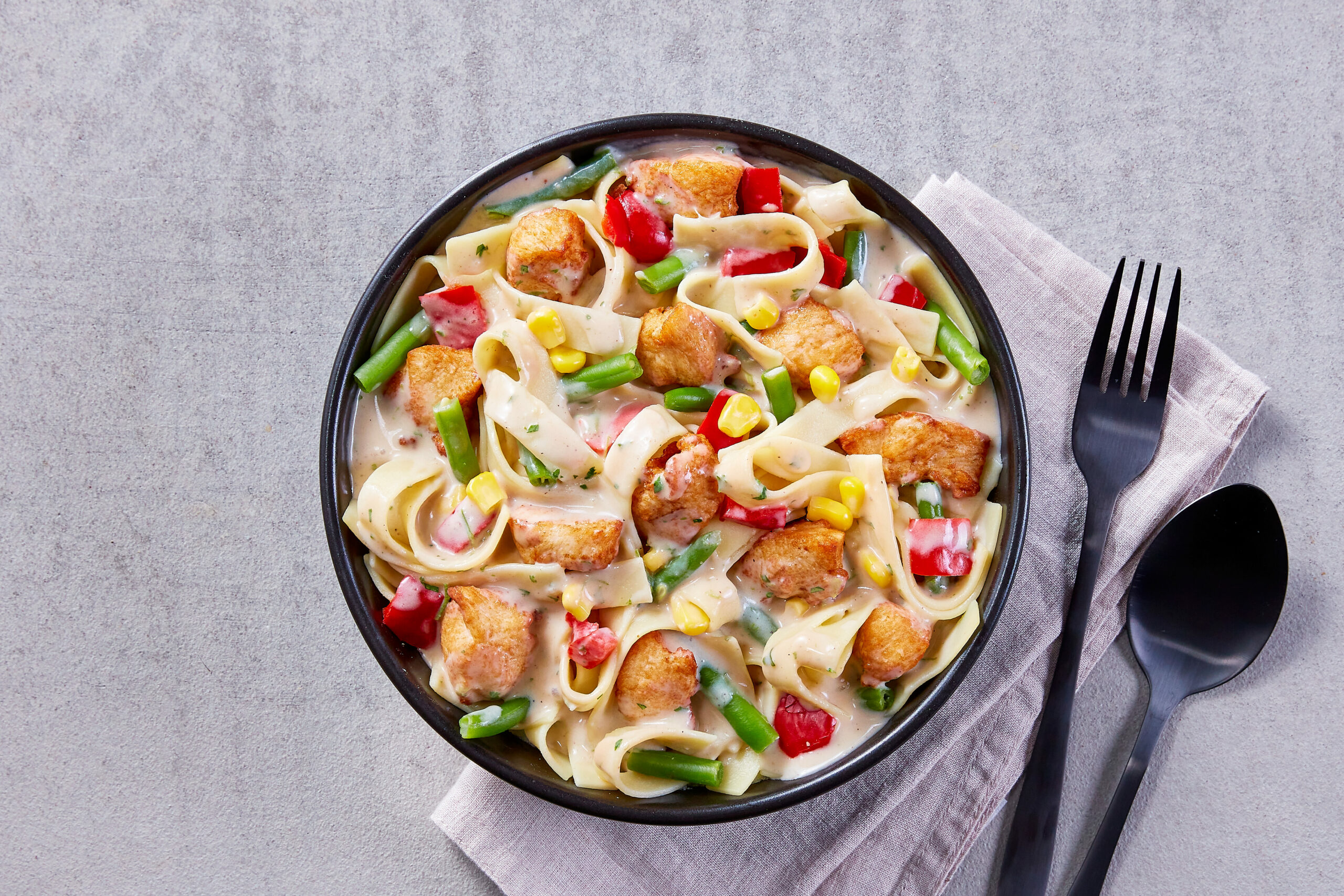 Eine Schüssel cremige Pasta mit gegrilltem Hähnchen, FRoSTA-Bohnen, FRoSTA-Paprika und FRoSTA-Mais auf einem grauen Untergrund mit Besteck.