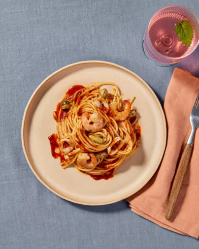 Ein Teller Spaghetti Frutti di Mare mit FRoSTA-Garnelen, Tomatensoße, einer Serviette in Pfirsichfarbe, einer Gabel und einem rosa Getränk mit Minzblatt.