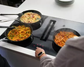 Zwei Personen kochen drei Pastagerichte; verschiedene Nudelsorten und bunte Soßen in Pfannen, einer trägt eine Smartwatch, im Hintergrund sind Teller zu sehen.